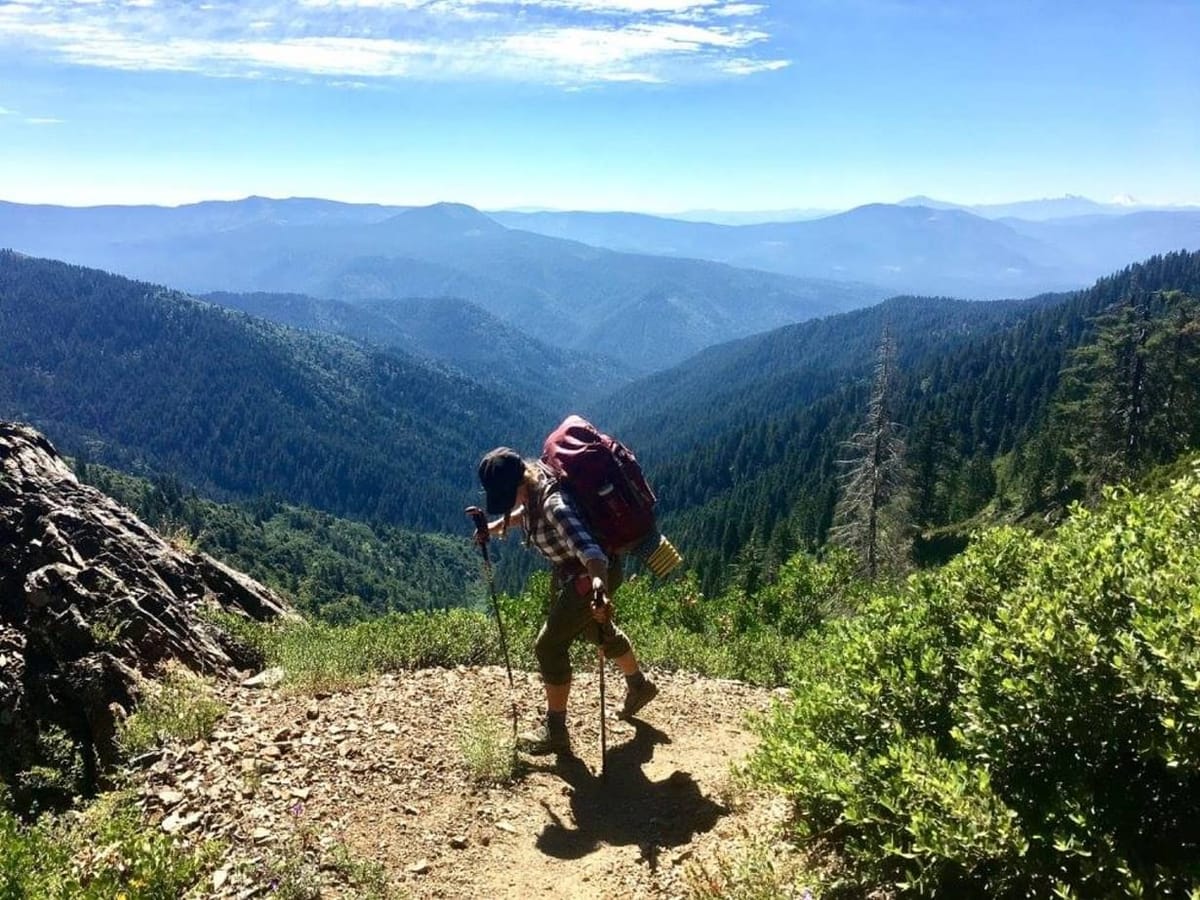 A backpacker on a sunny trail overlooking forested mountain ridges.