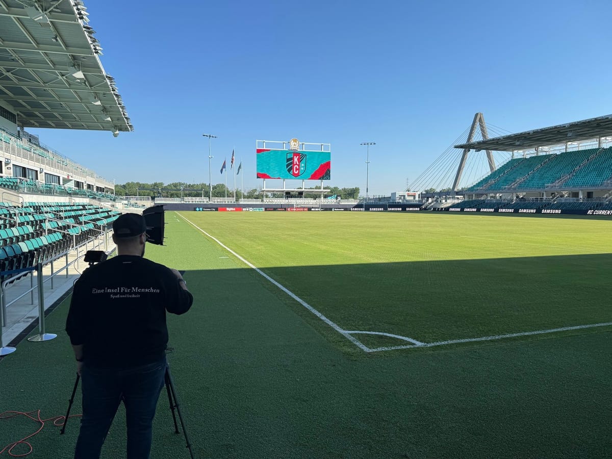 A camera operator filming from the sideline of a soccer stadium.